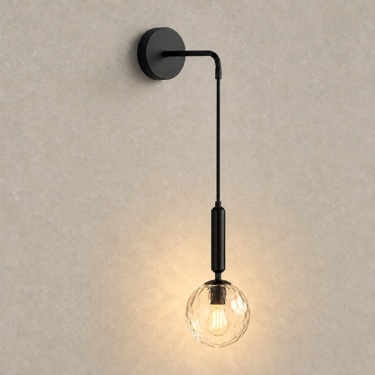 Orlamen black wall sconce switched on, the clear glass globe glowing on greige plaster