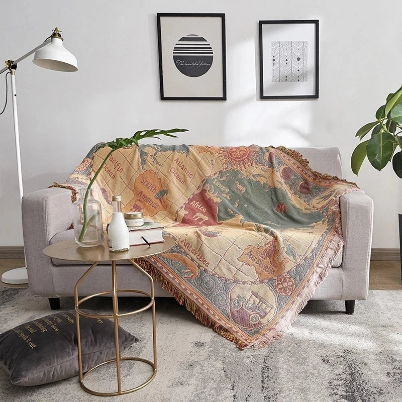 Saloveri world map throw blanket draped over a gray sofa, showing its vintage map design and fringed edges.