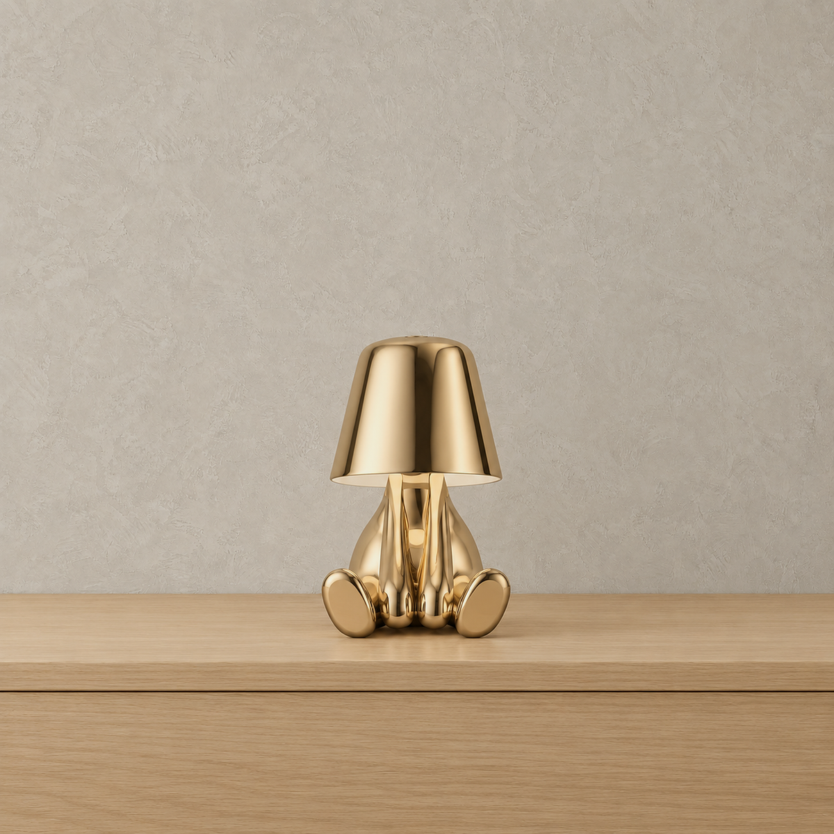 Sarovel gold rechargeable table lamp standing unlit against plaster, showing the polished seated figure and shade