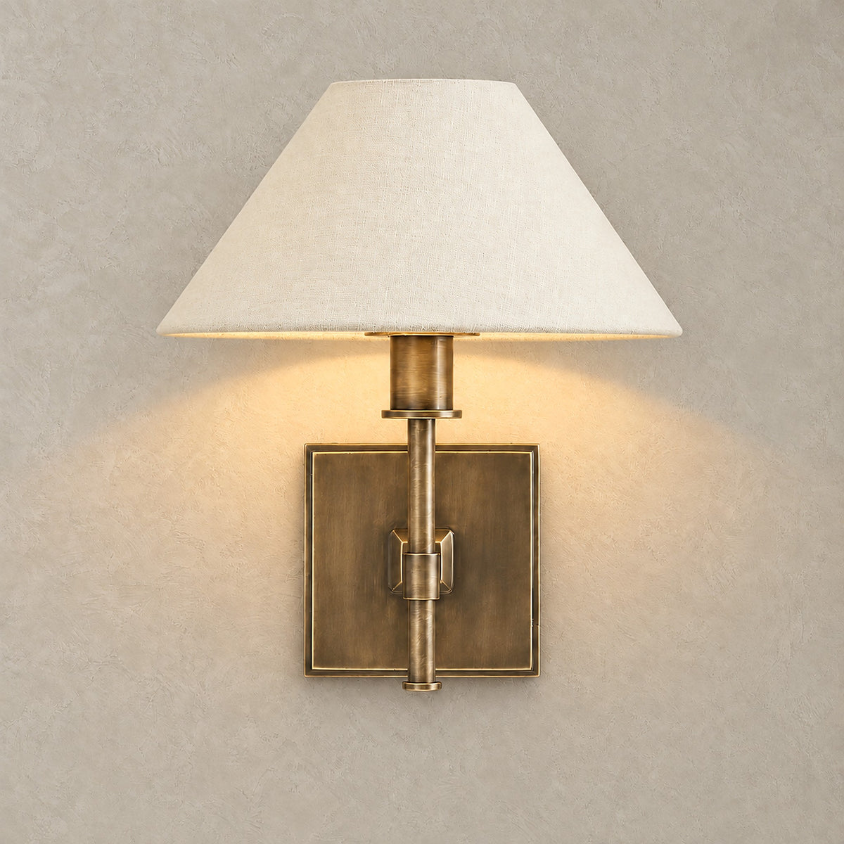 Ciralmo gold wall sconce with fabric shade lit against greige plaster