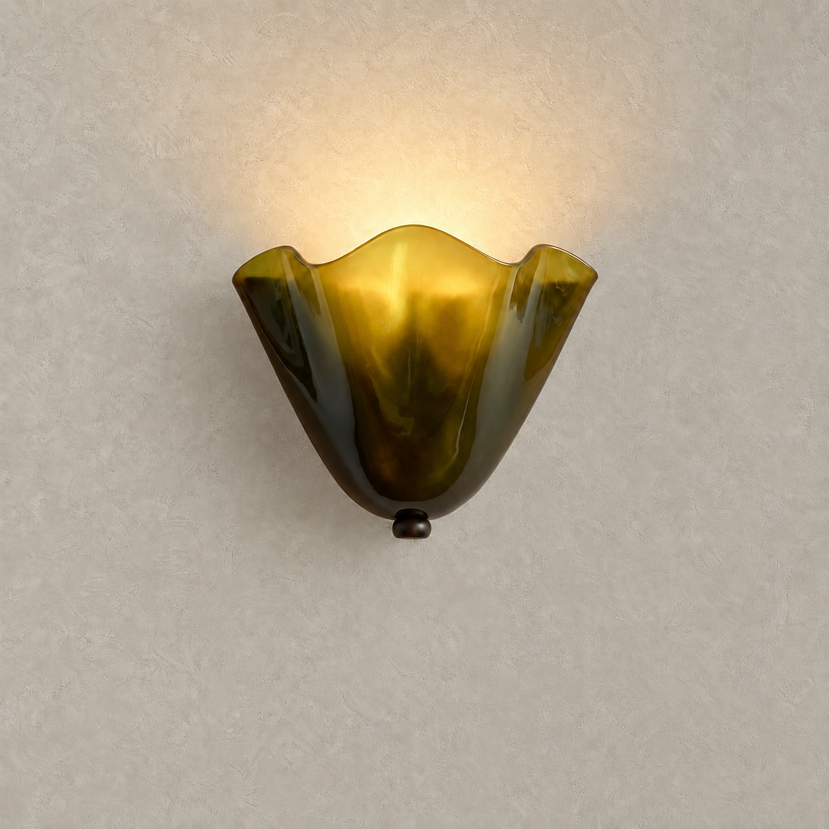 Ilsaren green wall sconce lit from inside its folded glass against warm greige plaster