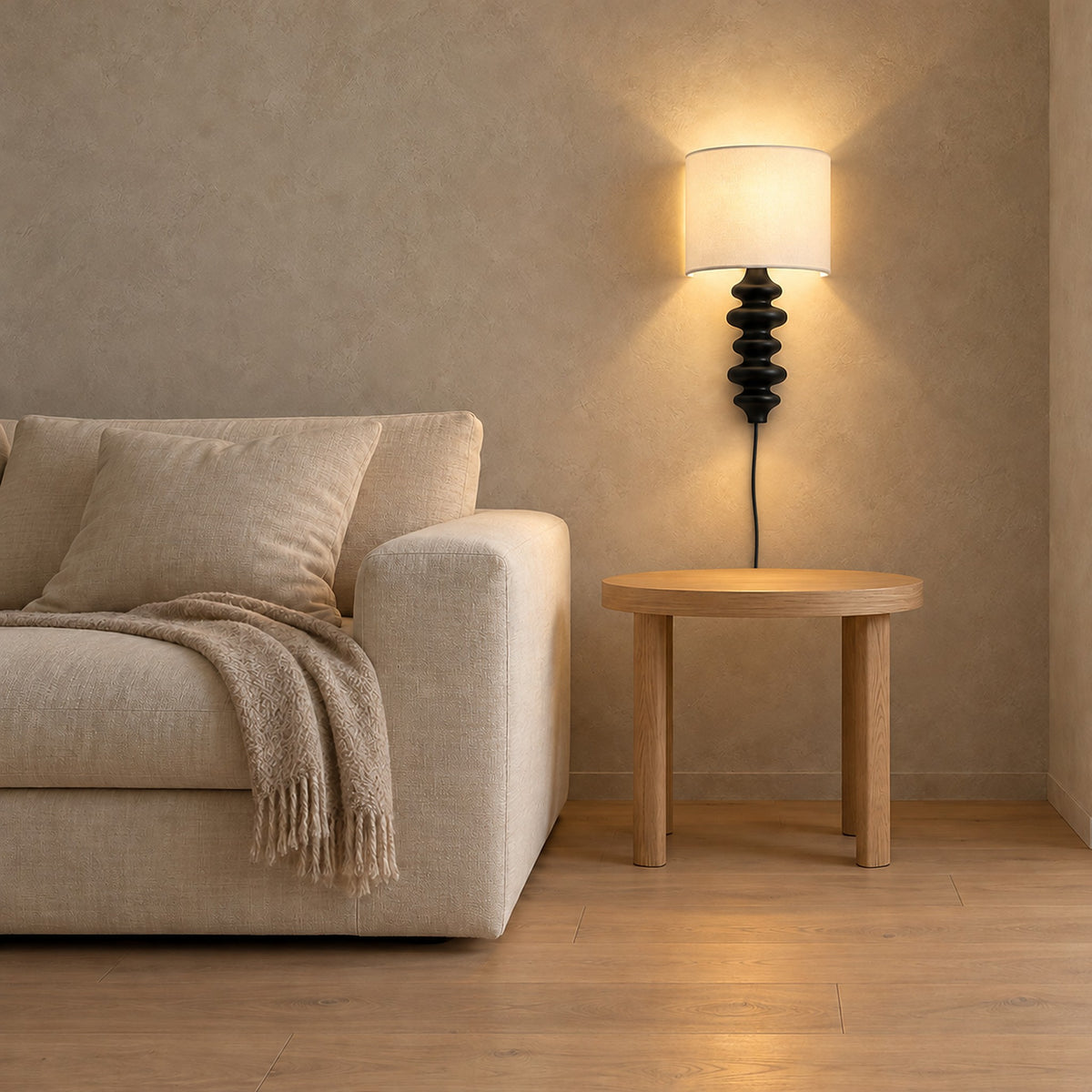 Nalimor Black wall lamp with a light shade, lit above an empty wooden side table beside a cream sofa.