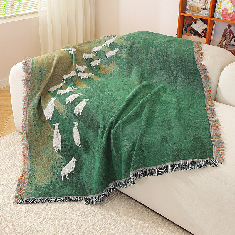 Timarno green woven sheep throw blanket draped over a white sofa, showing white sheep motifs and fringed edges.
