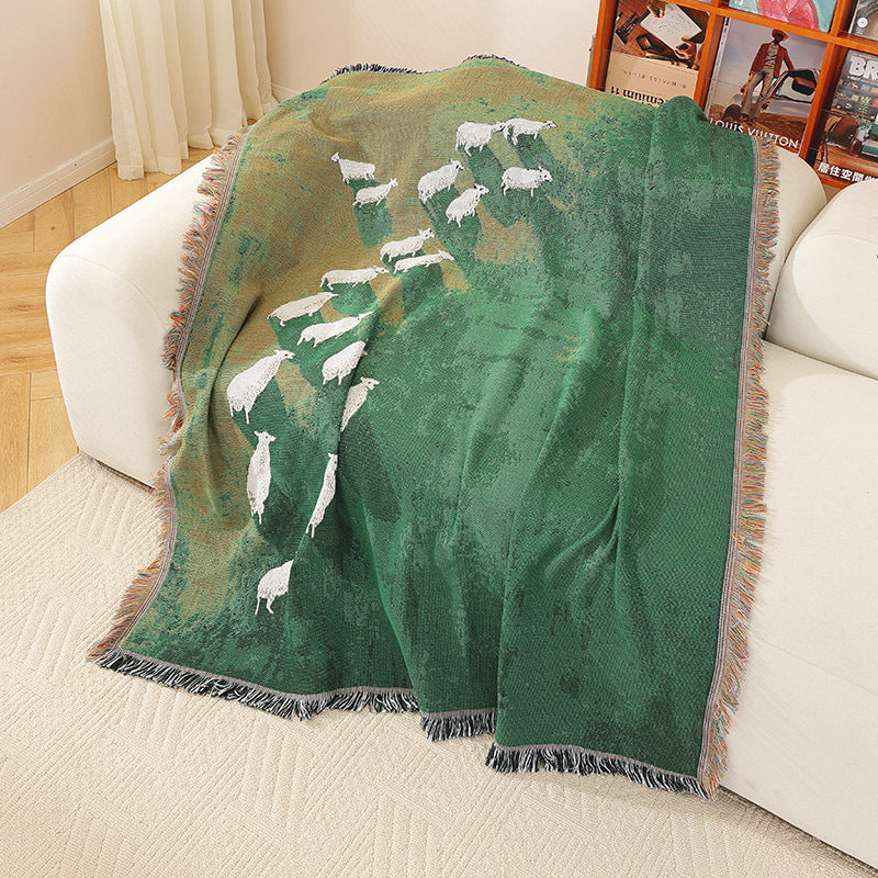 Timarno green woven sheep throw blanket draped over a cream sofa, showing white sheep motifs and fringed edges.