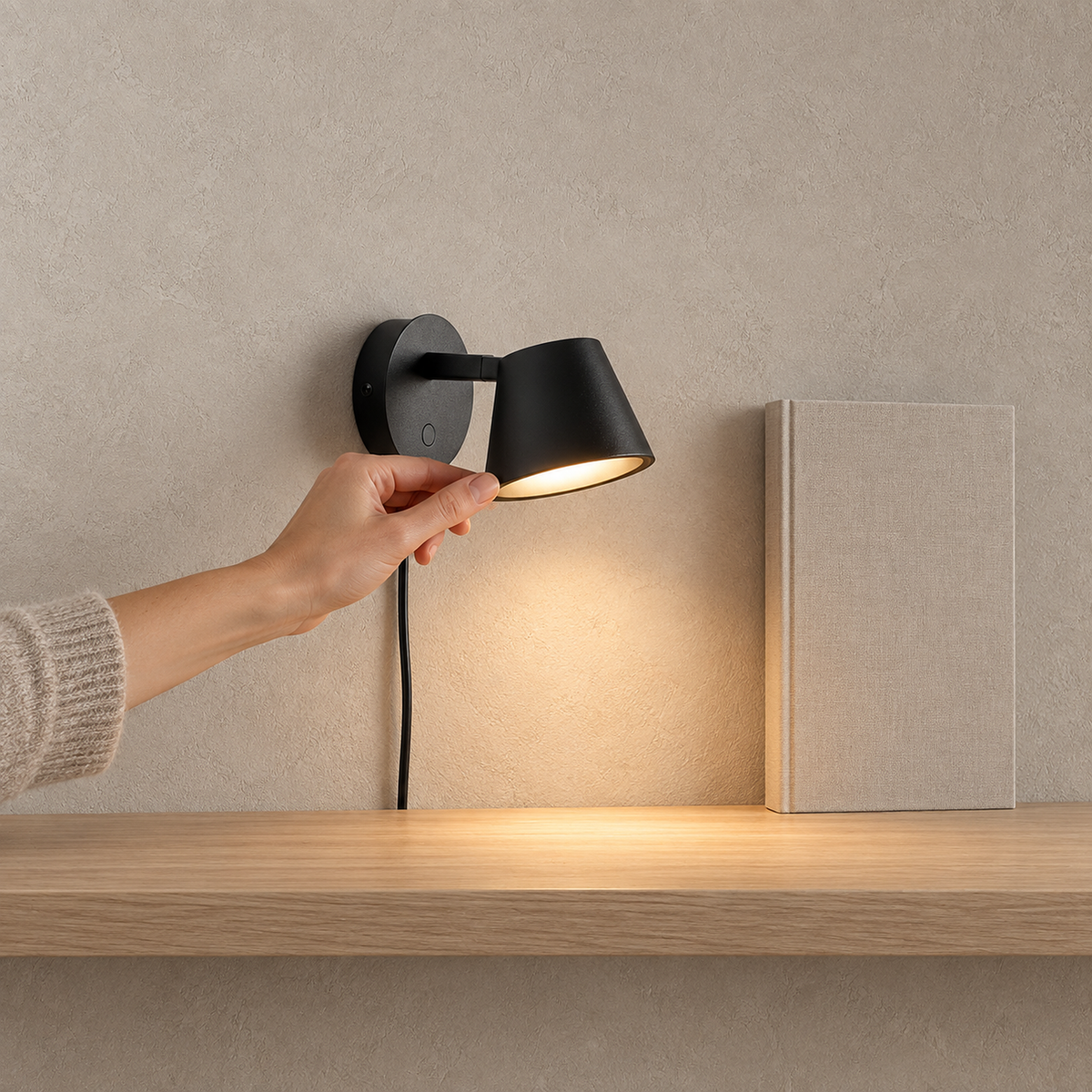 Tisello black adjustable wall sconce glowing as a hand rests lightly on the shade