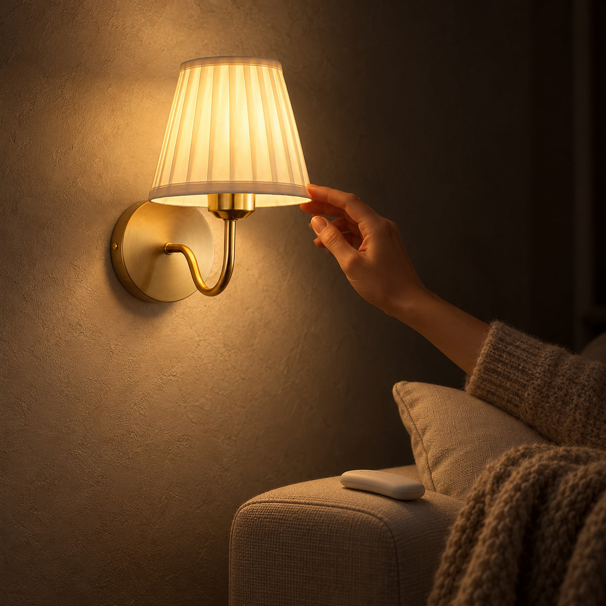 Tomirel gold wall lamp with a lit pale pleated shade beside an upholstered armchair, a hand reaching toward it